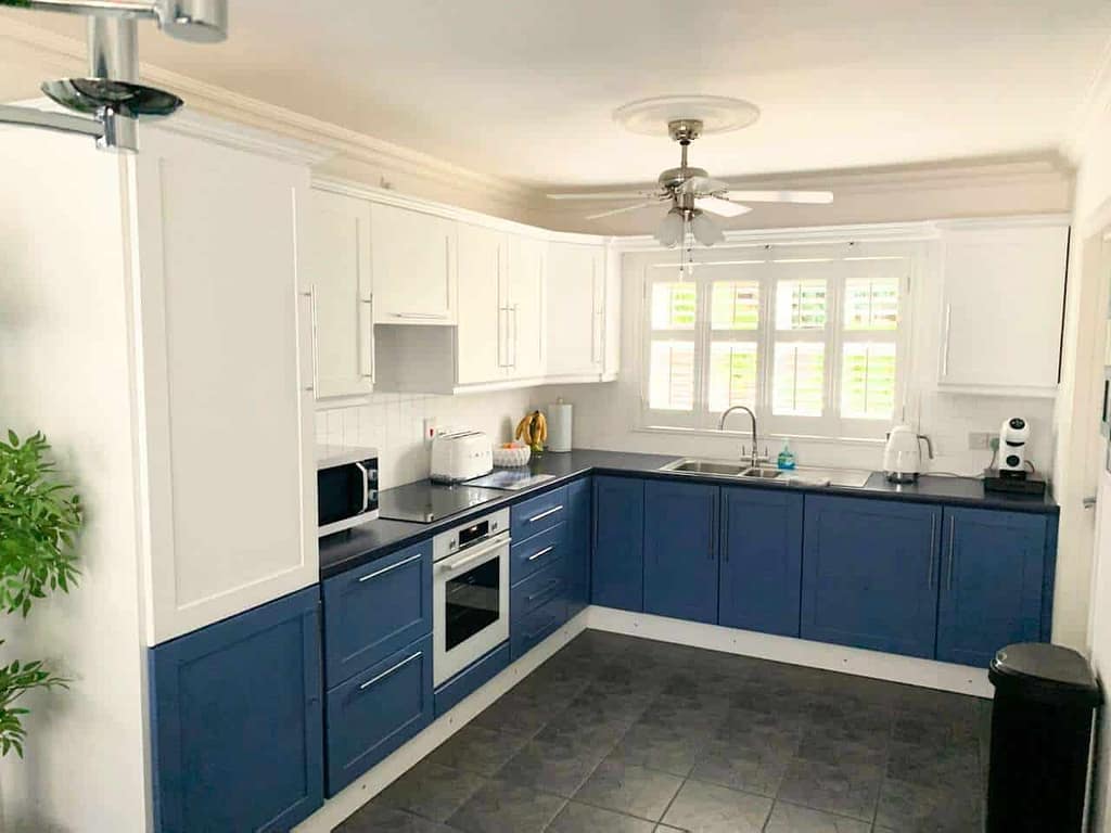 Bright kitchen interior with white upper cabinets and blue lower cabinets, modern appliances, large window with shutters, and ceiling fan, showcasing expert interior painting services in Portstewart.