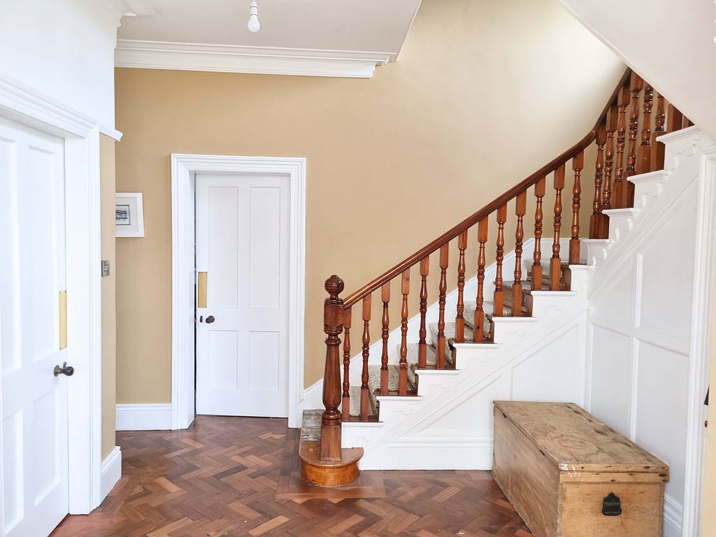 Bright interior hallway with freshly painted beige walls and white trim, featuring a wooden staircase and a vintage wooden storage chest, showcasing expert interior painting services in coleraine