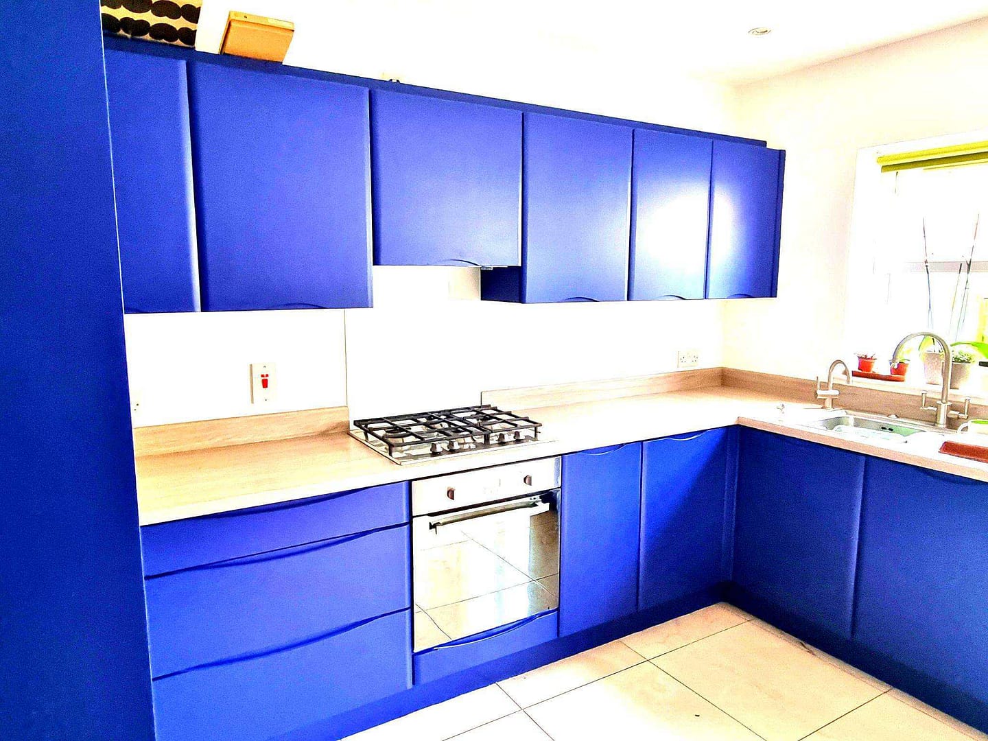Bright blue kitchen cupboards with wooden worktops, white walls and tiled flooring.